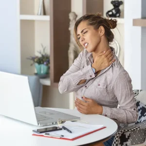 Mujer joven en el living de su hogar trabajando, contacta con su cuello y expresa una emoción de dolor