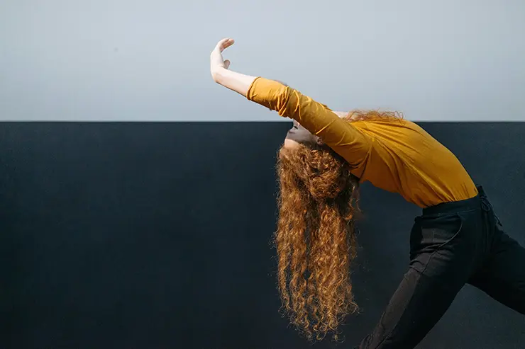 Mujer con rulos, sudadera mostaza arqueándose con los brazos extendidos hacia atrás danzando, 