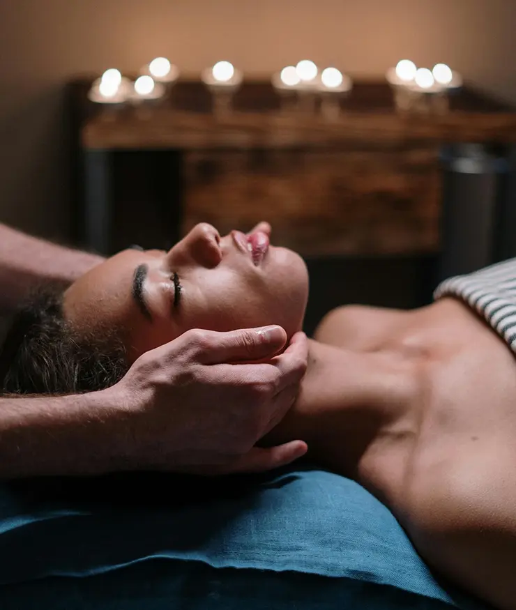 Mujer recostada recibiendo un tratamiento osteopático en el cuello. Detrás y de forma desenfocada hay unas velas encendidas.