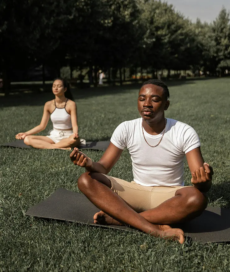 Una mujer y un hombre sentados sobre el pasto meditando, con los pies cruzados, manos apoyadas sobre rodillas y ojos cerrados.