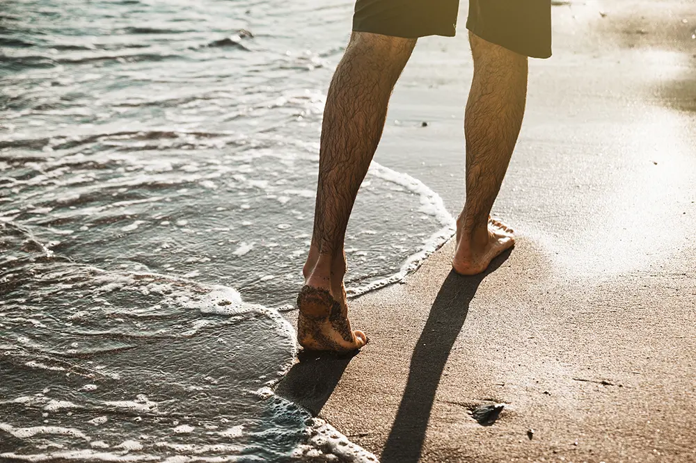 Hombre al que solamente de le ven las piernas que está caminando por la playa mientras el agua roza sus pies.