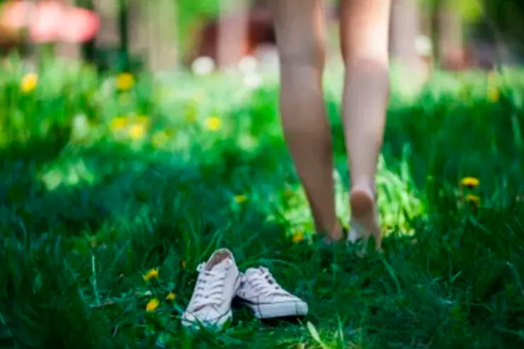 Piernas de una persona vistas desde atrás, las piernas están desenfocadas. La persona se encuentra descalza caminando sobre el césped.
En primer plano aparecen unas zapatillas.