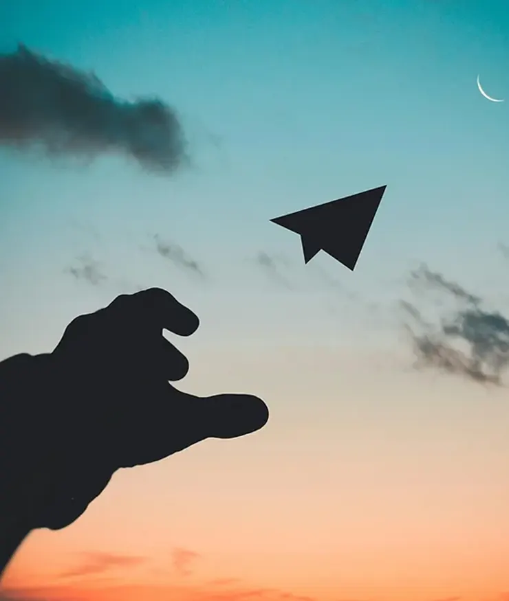 Mano de una persona lanzando un avión de papel al aire sobre un fondo de atardecer.