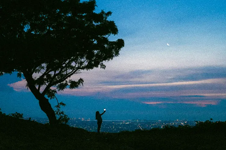 Persona de pie en la cima de una colina al atardecer, sosteniendo un teléfono que emite una luz. A la izquierda, un árbol solitario, y al fondo, una vista panorámica de una ciudad iluminada bajo un cielo azul y púrpura con una luna creciente visible.