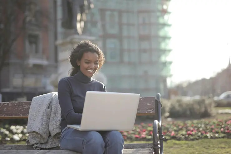 Mujer sentada en un banco de parque, sonriendo mientras usa una laptop. Lleva un suéter de cuello alto y tiene una chaqueta apoyada en el banco. Al fondo, un edificio urbano borroso y un jardín con flores iluminado por la luz del sol.