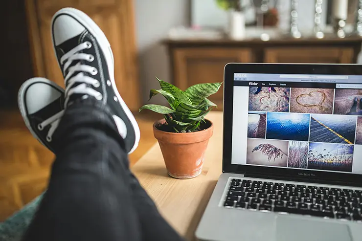 Persona relajada con los pies cruzados sobre una mesa, vistiendo zapatillas negras tipo Converse. Sobre la mesa hay una laptop mostrando una galería de imágenes y una maceta con una planta pequeña. Al fondo, muebles de madera desenfocados en un ambiente interior.