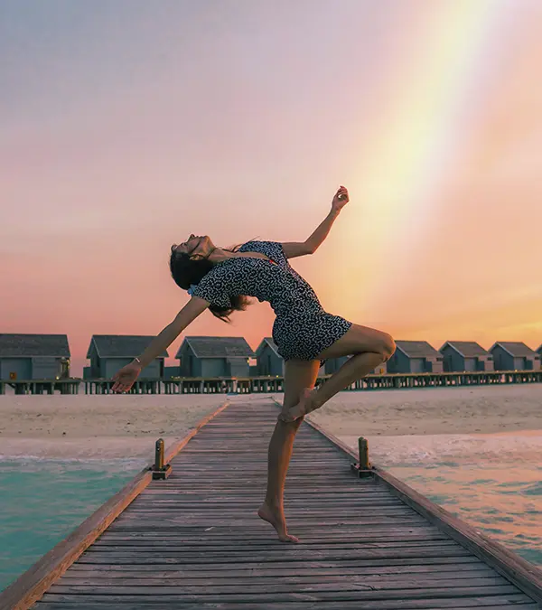 Un plano general muestra a una mujer delgada de piel clara, de aproximadamente 26 años, con cabello oscuro, equilibrándose con gracia sobre una pierna en un muelle de madera. Lleva un vestido corto con estampado. Su cuerpo se arquea hacia atrás, con los brazos extendidos, frente a un cielo al atardecer con tonos pastel. A lo lejos, una fila de pequeñas casas de playa idénticas se alinea en la orilla. El muelle se adentra en el agua turquesa, creando sensación de profundidad y tranquilidad. La imagen está bañada con luz suave y difusa, realzando la atmósfera pacífica.
