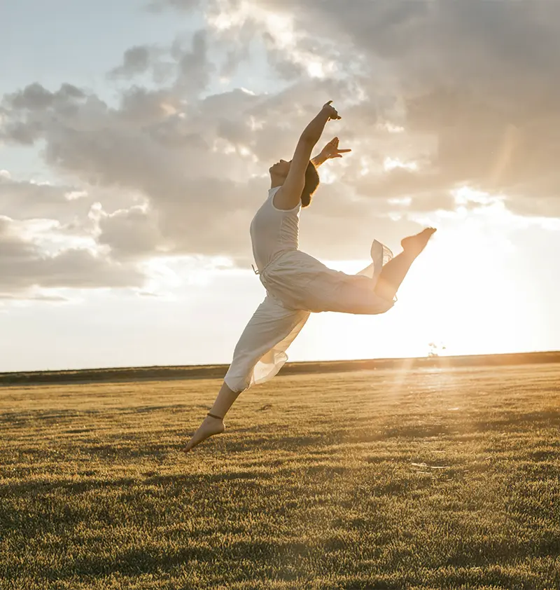 Un plano general muestra a una mujer blanca de 23 años en el aire, aparentemente bailando o saltando, frente a un campo y un cielo lleno de nubes. La mujer, vestida con un top sin mangas blanco y pantalones a juego, tiene los brazos levantados y las piernas extendidas, transmitiendo una sensación de libertad y movimiento. El campo está cubierto de pasto corto y seco, y el cielo lleno de nubes deja pasar la luz del sol, creando una iluminación cálida y dorada. La luz es suave y difusa, realzando el ambiente general de la imagen. La composición es simple pero efectiva, con la mujer como foco principal y el fondo proporcionando contexto y profundidad. La imagen tiene un aspecto natural, sin retoques, enfocándose en capturar un momento en el tiempo.