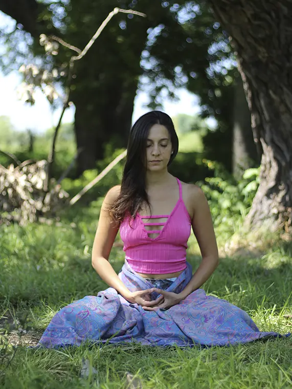 Un plano medio muestra a una mujer hispana de 24 años, con cabello largo y oscuro, vistiendo un top rosa y una falda azul con estampado, sentada en postura de meditación sobre el césped en un bosque. La iluminación es suave y difusa, creando una atmósfera tranquila.