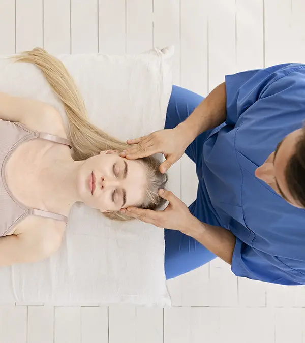 Plano cenital de una mujer blanca de 22 años, con piel clara y cabello rubio largo, recostada sobre una almohada blanca, recibiendo un masaje en la cabeza de un hombre con uniforme médico azul. La mujer lleva un top sin mangas rosa claro. El hombre tiene cabello oscuro y piel morena. El fondo es un piso de madera blanca. La iluminación es suave y difusa.