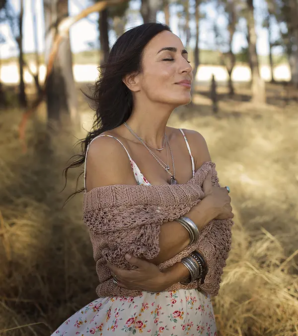 Un plano medio muestra a una mujer hispana de 42 años, con piel bronceada, cabello largo castaño oscuro y ojos cerrados, de pie en un campo con árboles de fondo. Viste un vestido blanco con estampado floral y un chal tejido color marrón claro sobre los hombros. Sus brazos están cruzados al frente y lleva varios collares y pulseras de plata. La iluminación es suave y difusa, creando una atmósfera cálida y natural. La composición sigue la regla de los tercios, con la mujer ligeramente desplazada del centro. El estilo general recuerda a una fotografía vintage con una textura ligeramente granulada.